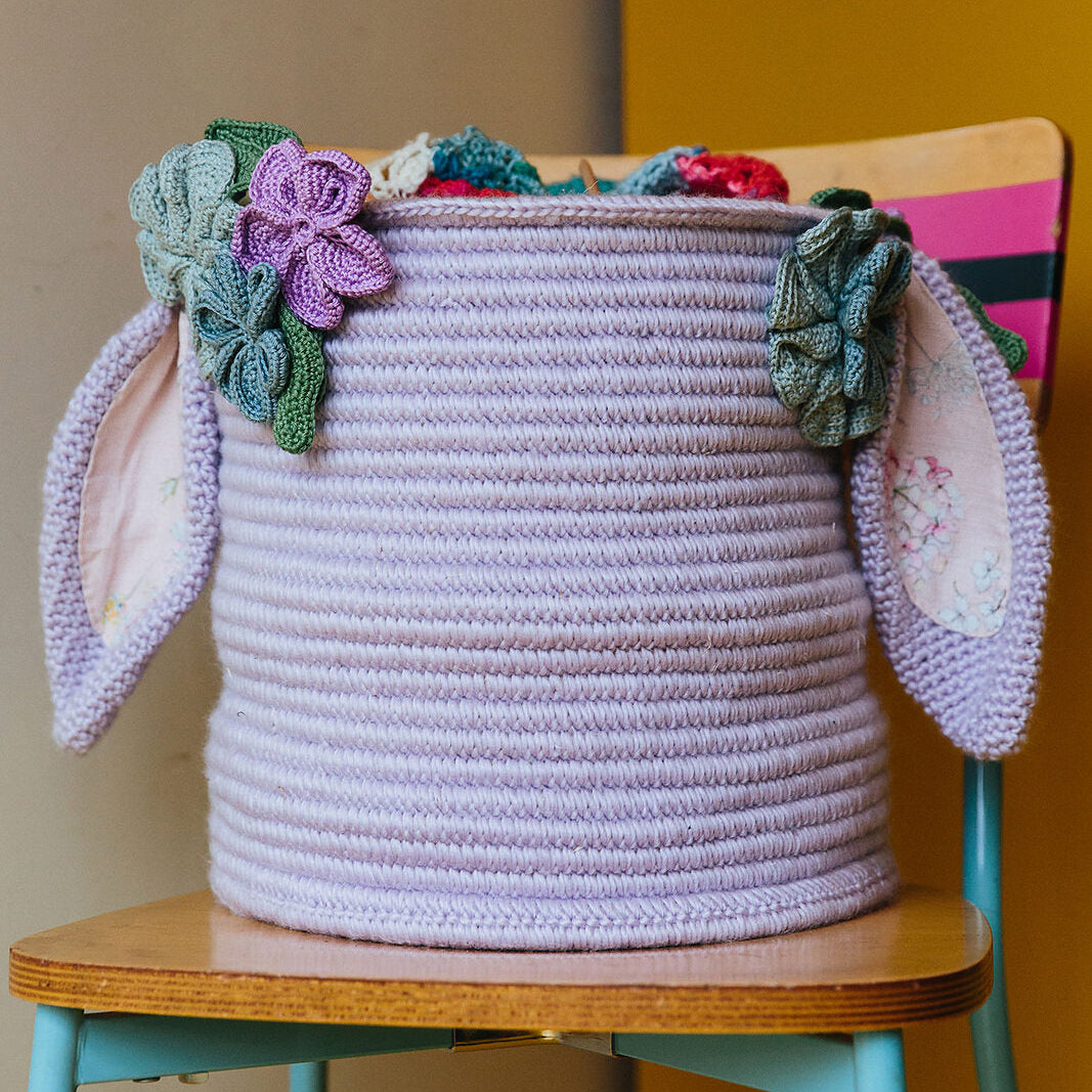 Decorative crocheted basket with floral embellishments and bunny ears on a chair against a yellow wall.